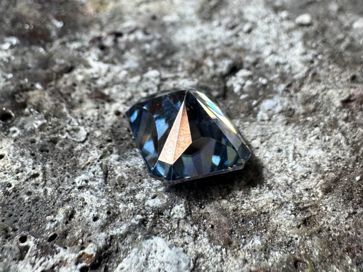 Close-up of the blue eBay stone tilted on its side. A pinkish copper-colored reflection flashes off one pavilion facet, revealing the coating.
