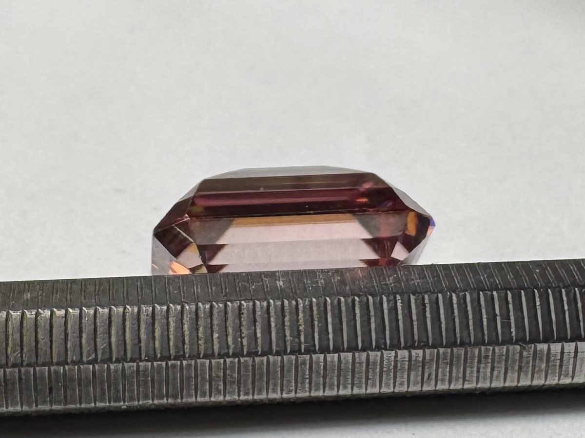 A pink eBay 'lab diamond' seen in profile, resting on a bench file. The pavilion below the girdle is coppery pink from the coating while the crown and table above it are clear.