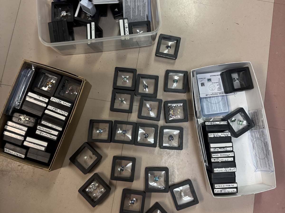 Dozens of loose stones in black display boxes spread across a floor beside two boxes of paperwork and labeled stone parcels. All were sold on eBay as lab grown diamonds.
